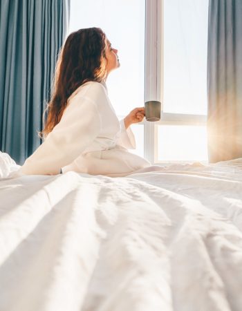 Pretty positive woman with long hair drinks water and sitting at bed in sunny lazy morning.